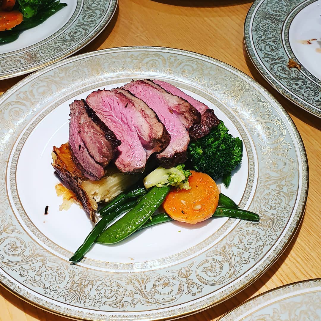 Sliced medium-rare beef rests on a decorative plate, accompanied by vibrant broccoli, green beans, and glazed carrots, all in warm ambient light.