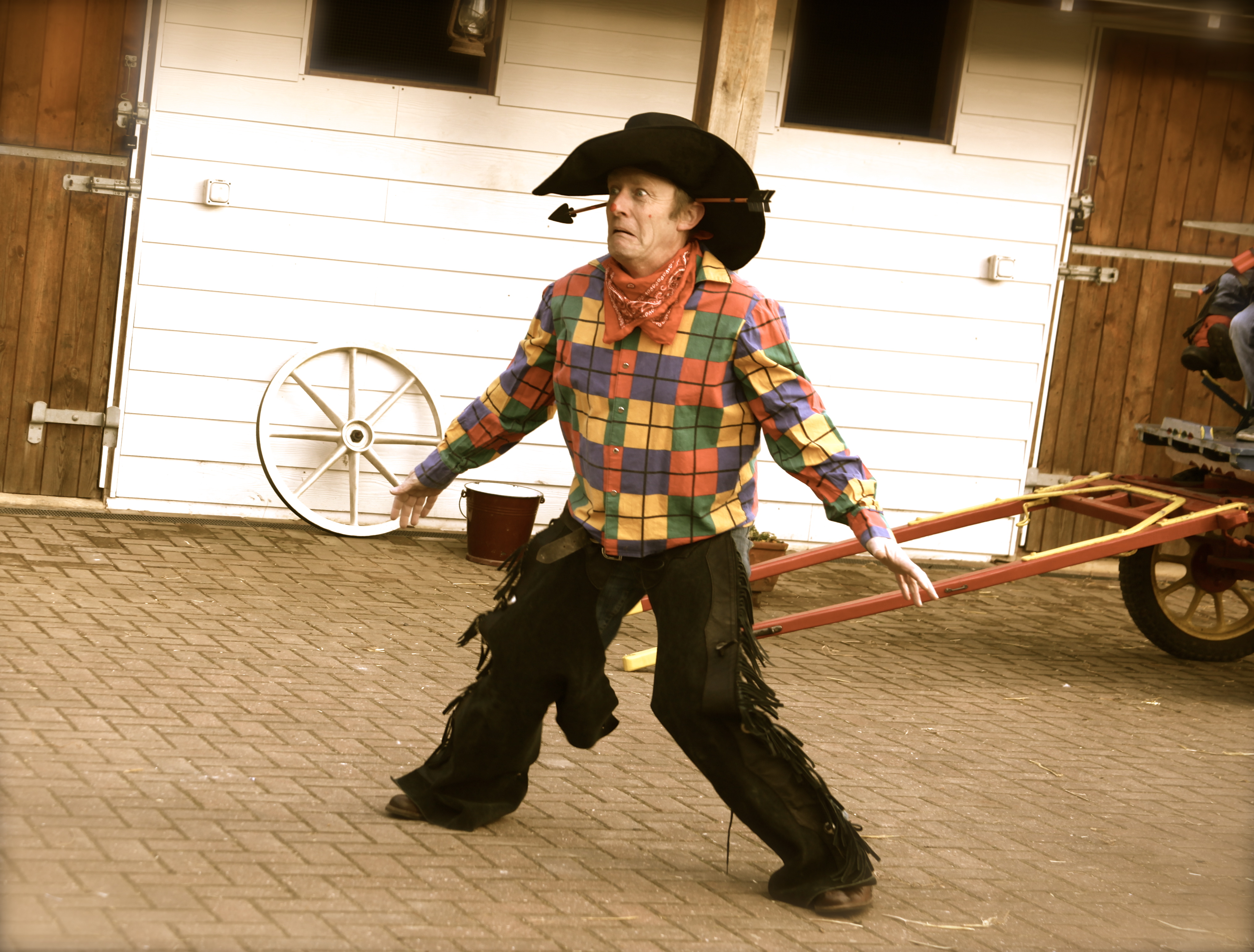 A lively performer in a checkered shirt and fringed pants entertains in a rustic outdoor setting, exuding vibrant, playful energy.