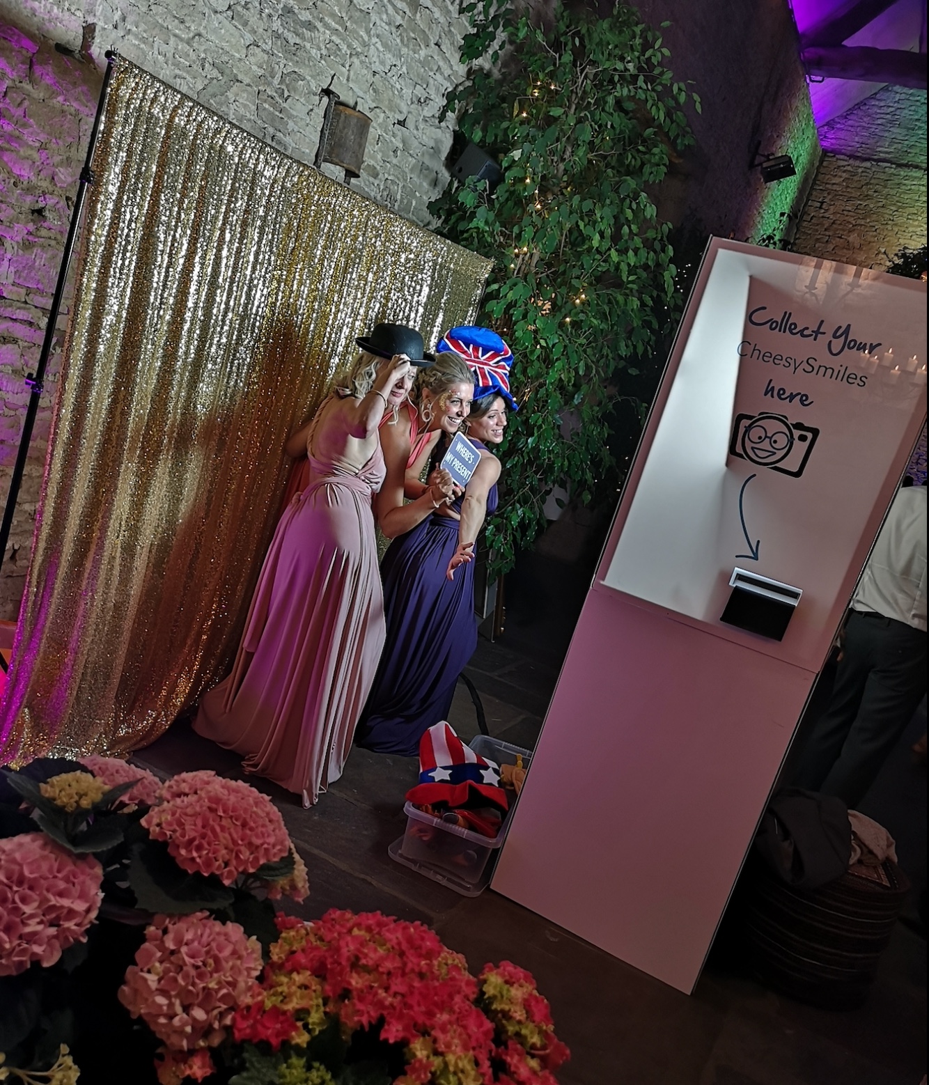 Three women pose playfully before a sparkling gold backdrop, surrounded by vibrant pink flowers, under soft, colorful lighting for a fun event.