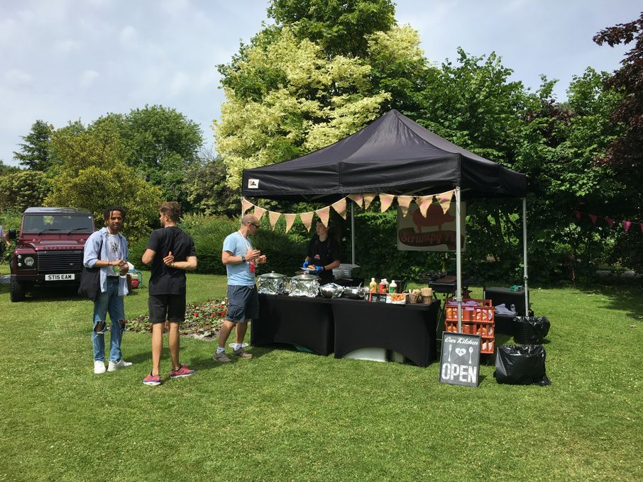 A black tent displays a vibrant buffet with neatly arranged silver trays and colorful sauces; sunlight creates a lively outdoor garden atmosphere.
