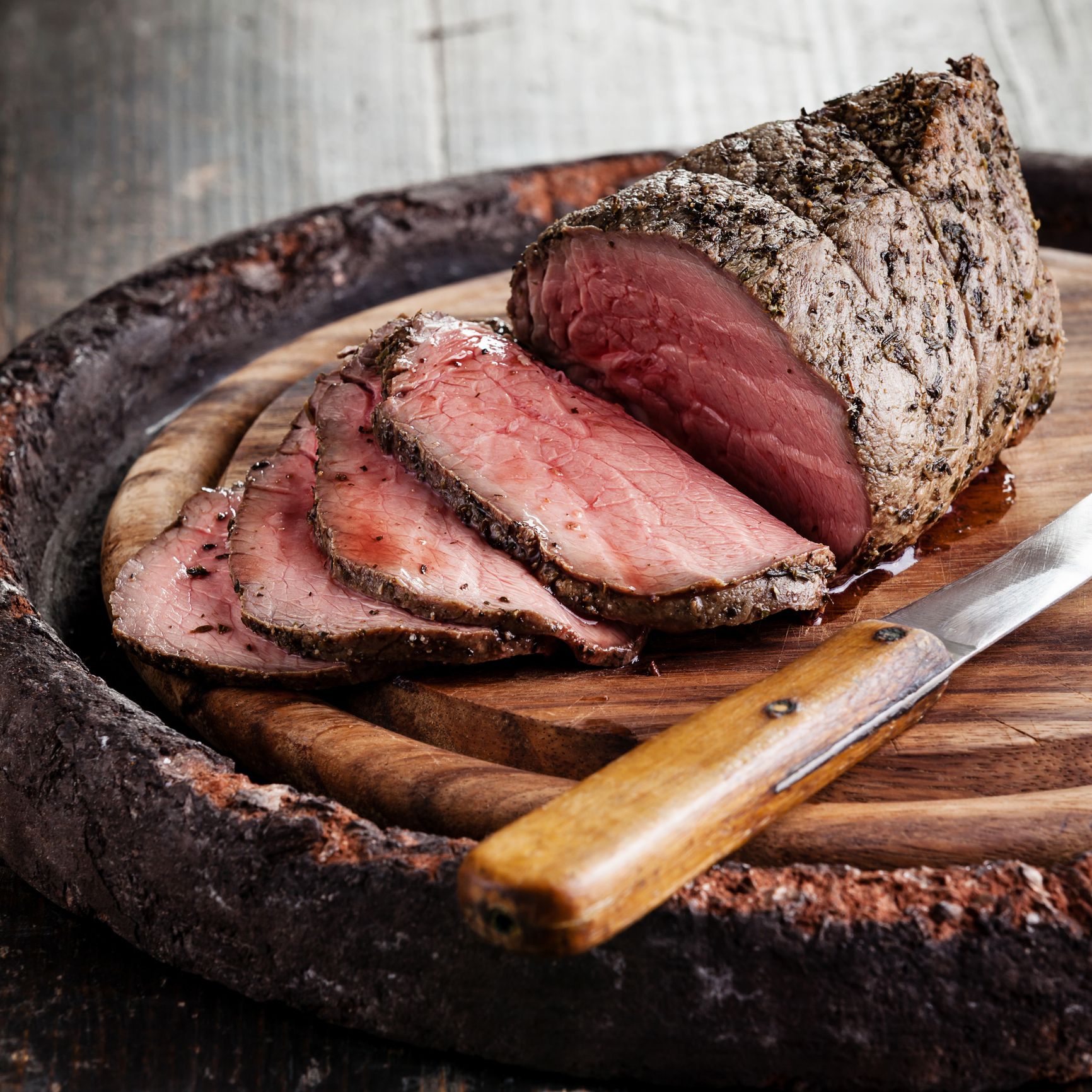 Juicy, herb-crusted roast beef sliced on a rustic wooden platter, paired with a polished knife, evokes a warm, inviting atmosphere perfect for catering.
