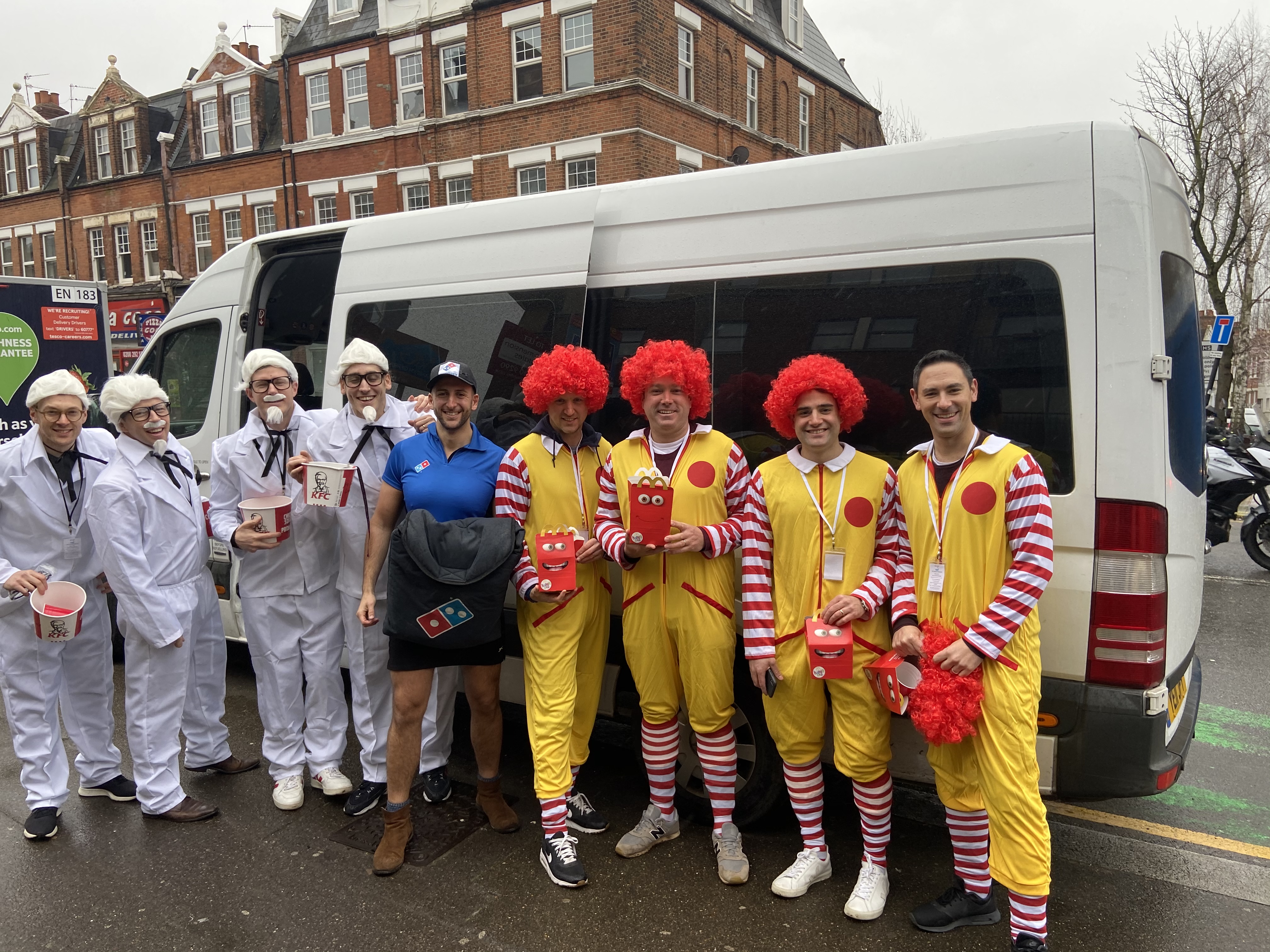 A lively group in comical fast-food costumes poses cheerfully beside a white van on a rainy street, showcasing vibrant colors and fun energy.