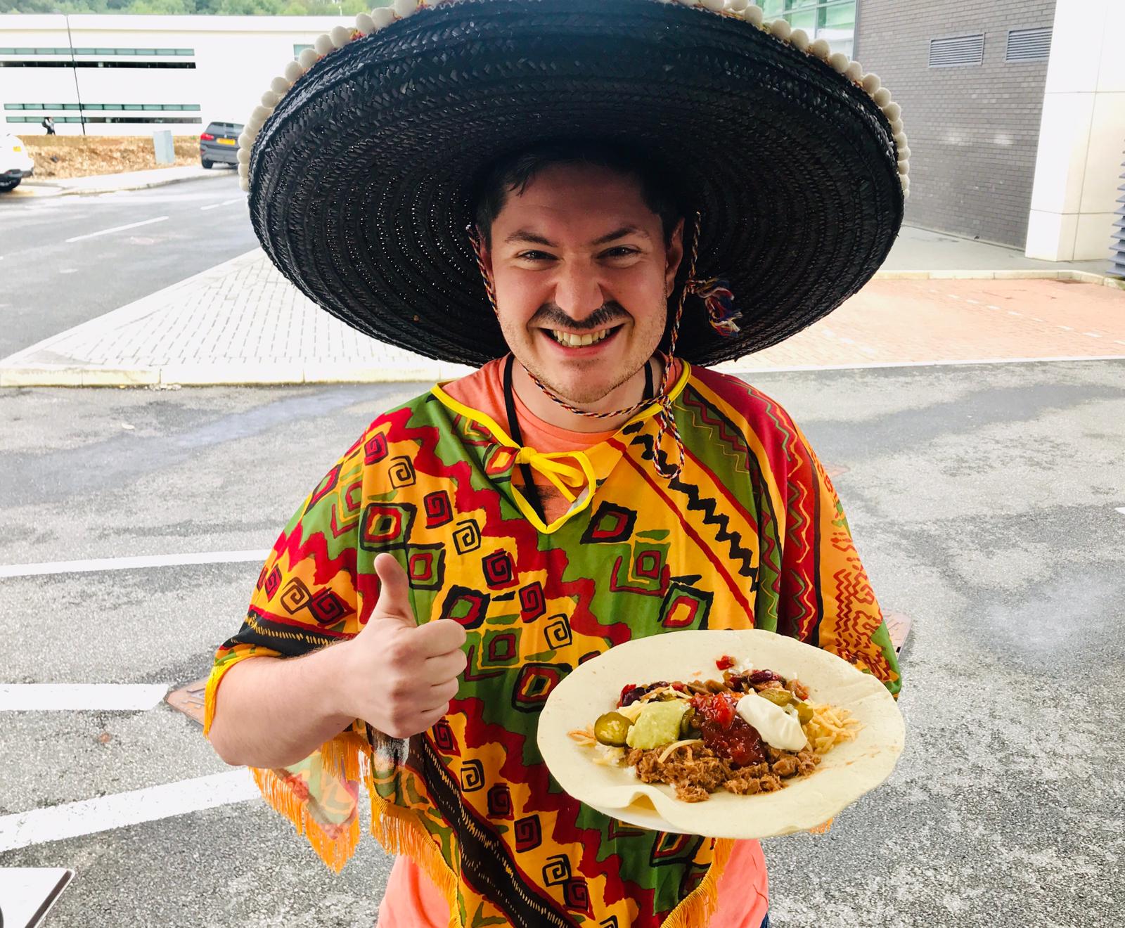 A cheerful man in a vibrant poncho and wide-brimmed hat holds a plate of colorful taco fillings, including salsa and guacamole, outdoors.