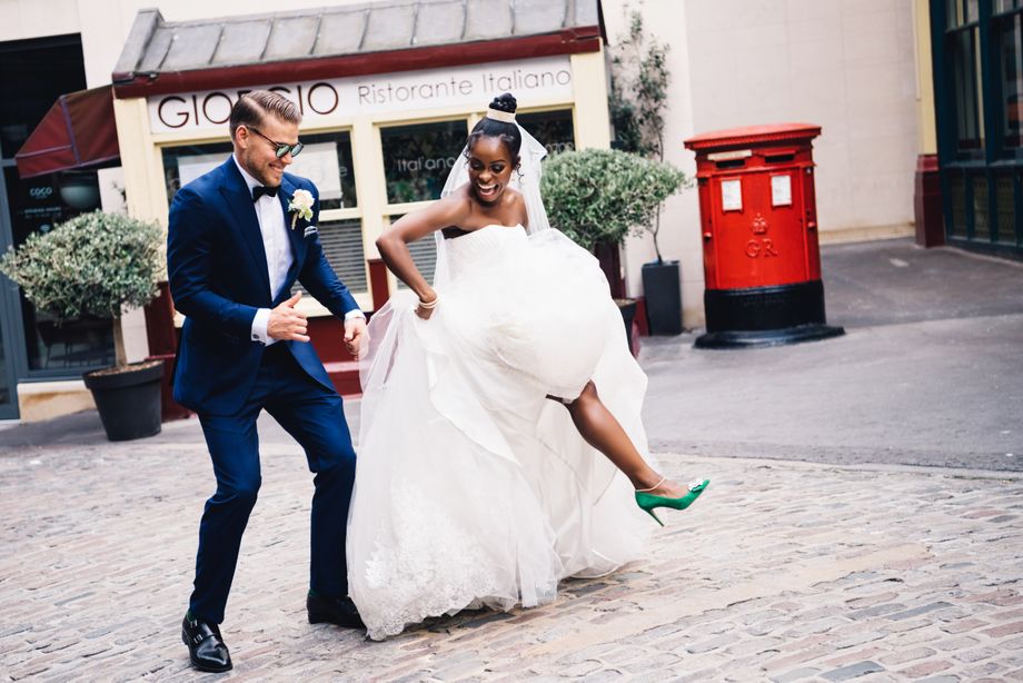 A joyful couple dances on cobblestone, surrounded by lush greenery and a vibrant red postbox, exuding festive wedding vibes in daylight.