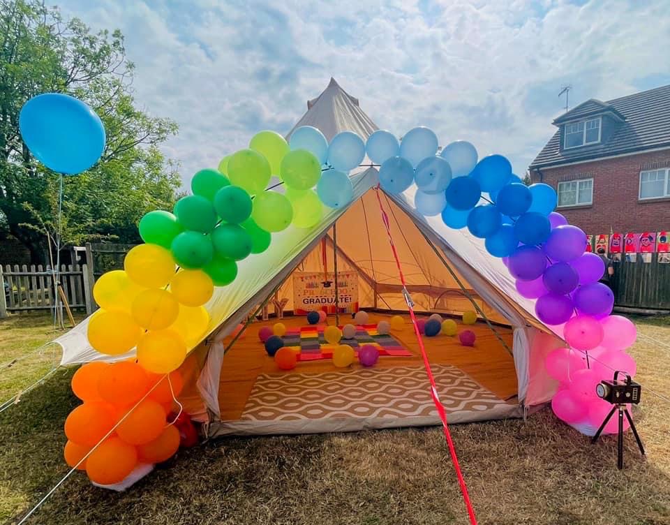 A festive tent decorated with a vibrant balloon arch in rainbow colors, set in bright daylight, featuring a cheerful atmosphere for a celebration.
