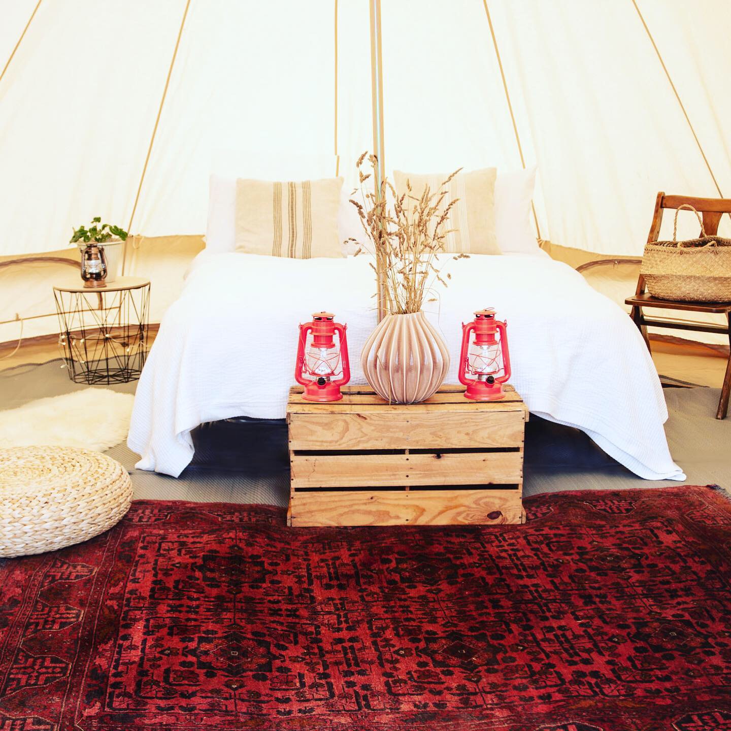 A cozy glamping setup features a white bedspread on a wooden crate, flanked by red lanterns, with a vintage rug and potted plants for warmth.
