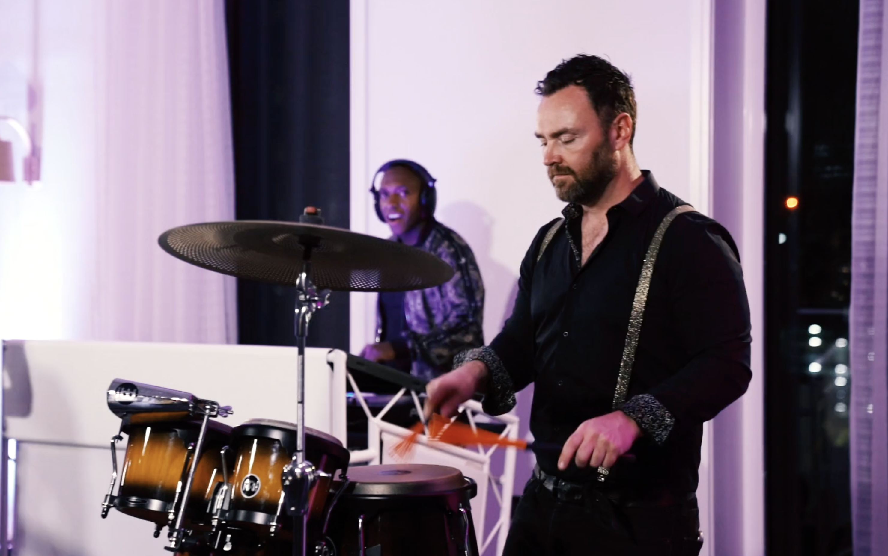 A drummer in sleek black with silver suspenders plays energetically while a smiling keyboardist in a sparkly jacket joins the rhythm.