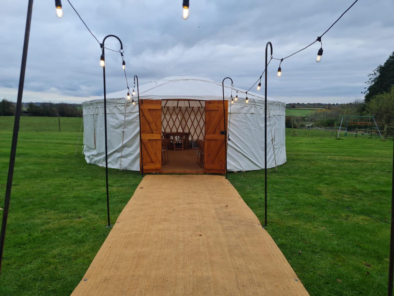 A charming yurt with wooden doors and twinkling string lights welcomes guests along a cozy path, set against a serene green field.