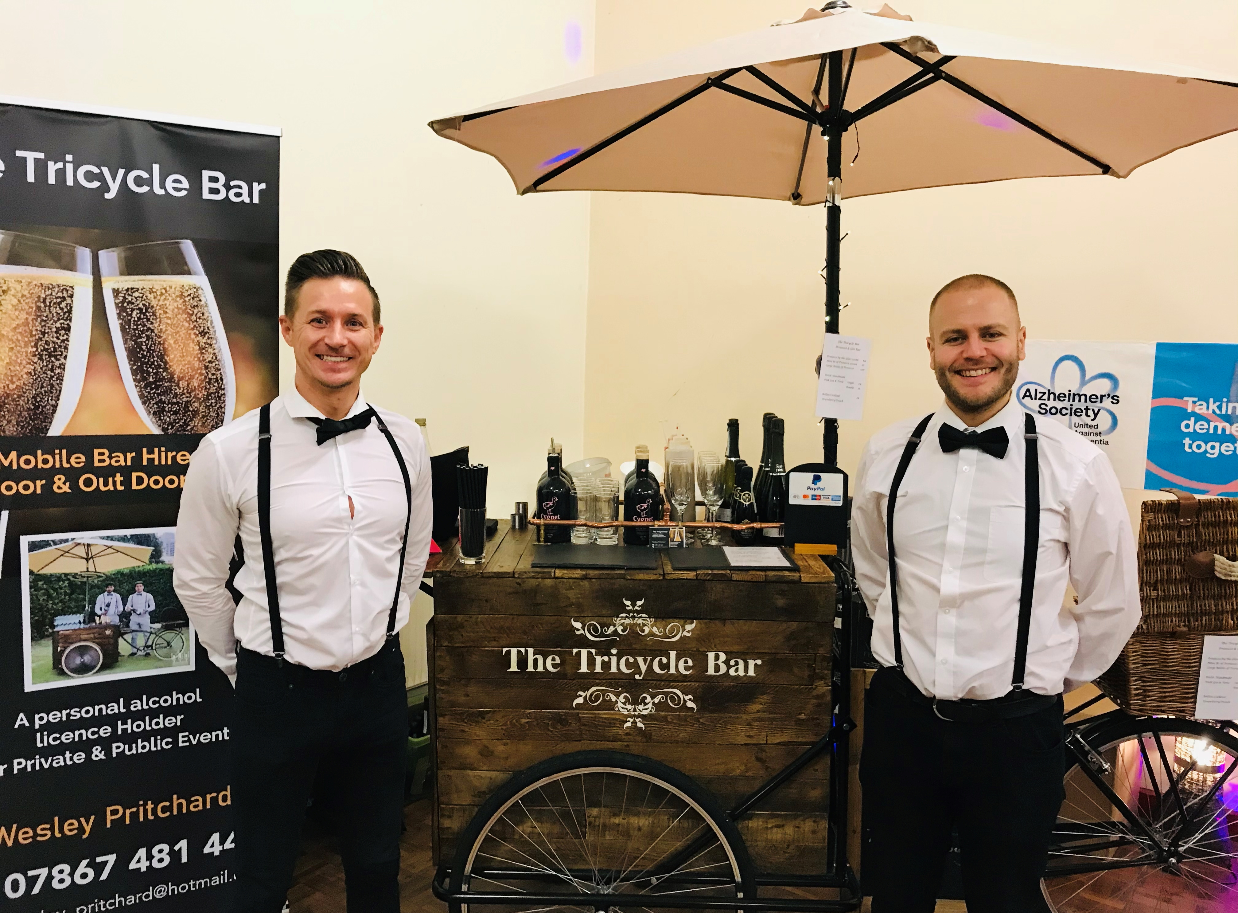 The Tricycle Bar features a rustic wooden mobile bar with polished glassware, manned by two formally dressed bartenders under a beige umbrella, showcasing service quality.
