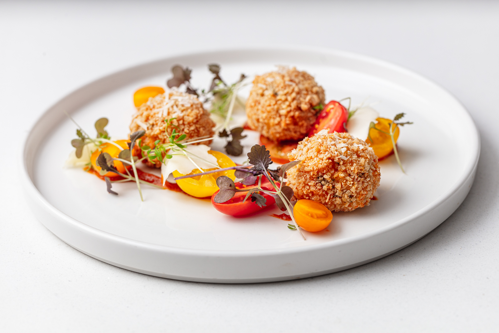 Crispy breaded balls sit elegantly on a smooth white plate, garnished with vibrant yellow and red cherry tomatoes, microgreens, and drizzled sauce, offering a refined atmosphere for gourmet catering.