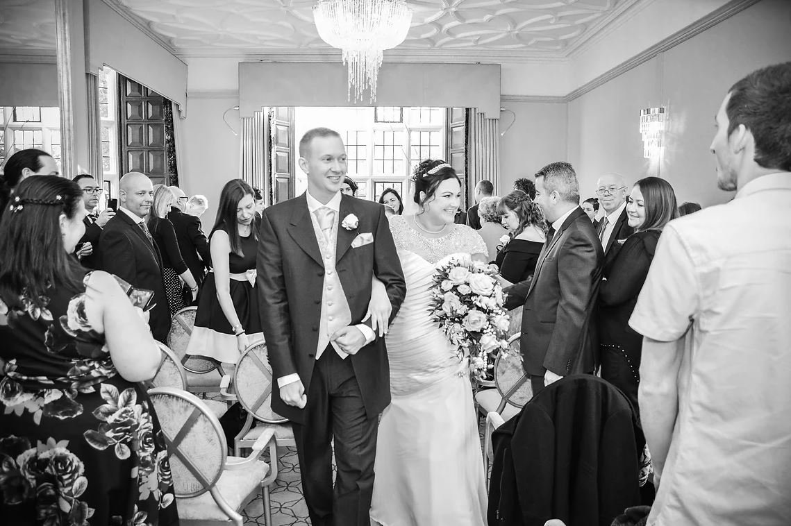 A joyful couple exits a ceremony in an elegant venue with decorative chandeliers, surrounded by guests in formal attire, exuding celebration.