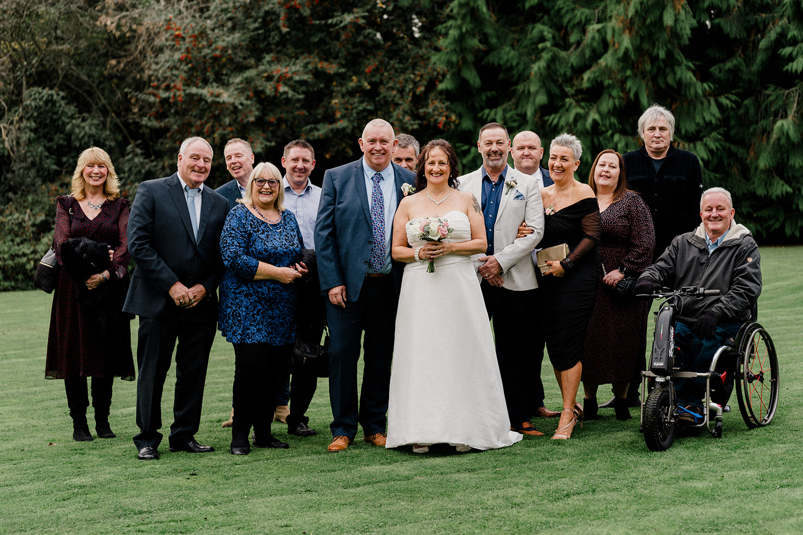 A joyful wedding party poses on lush green grass, featuring a bride in a flowing white gown and diverse guests in varying attire.