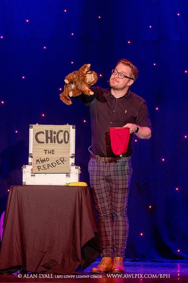 A performer showcases a puppet while standing beside a vintage-style box labeled "Chico, the Mind Reader," under vibrant stage lights, exuding a playful atmosphere.