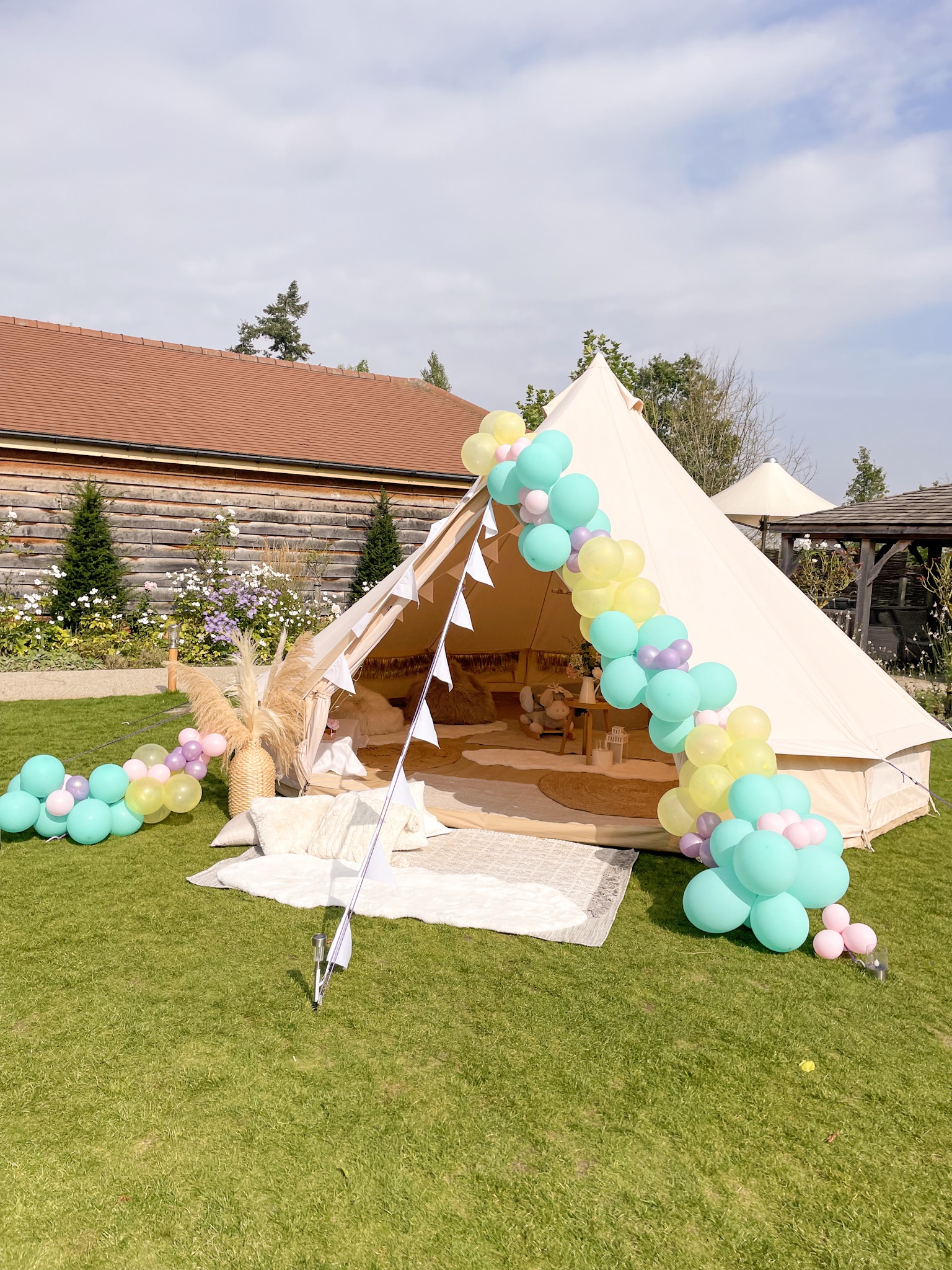 A cozy tent adorned with pastel balloons spills onto lush green grass, creating a whimsical space perfect for outdoor gatherings, bathed in soft daylight.
