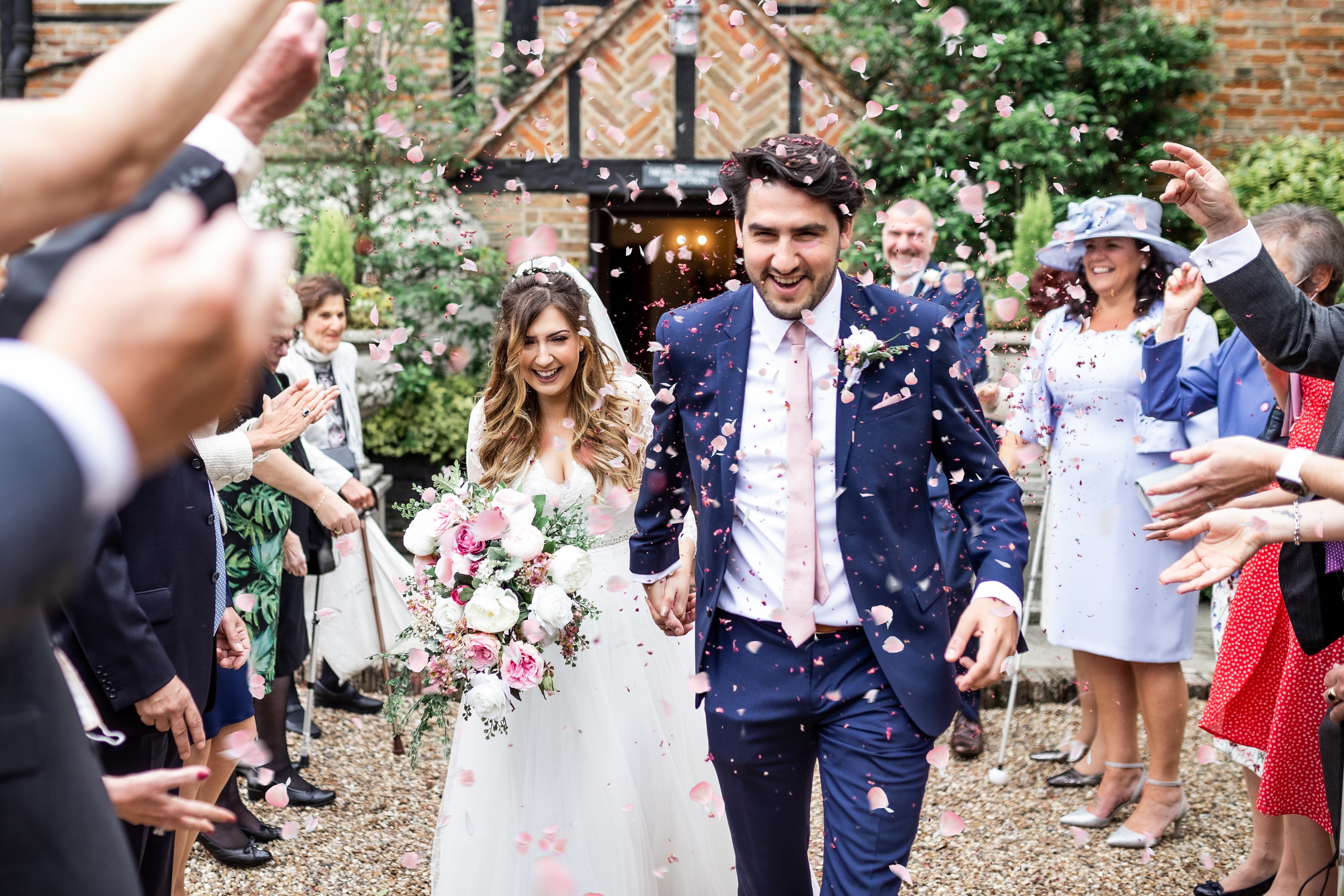 A joyful couple exits a charming venue, surrounded by cheering guests tossing pink rose petals, creating a vibrant, festive ambiance.