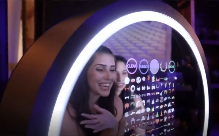 Two women smile in front of a sleek, illuminated touch screen displaying vibrant emojis and icons, creating a fun, engaging atmosphere.