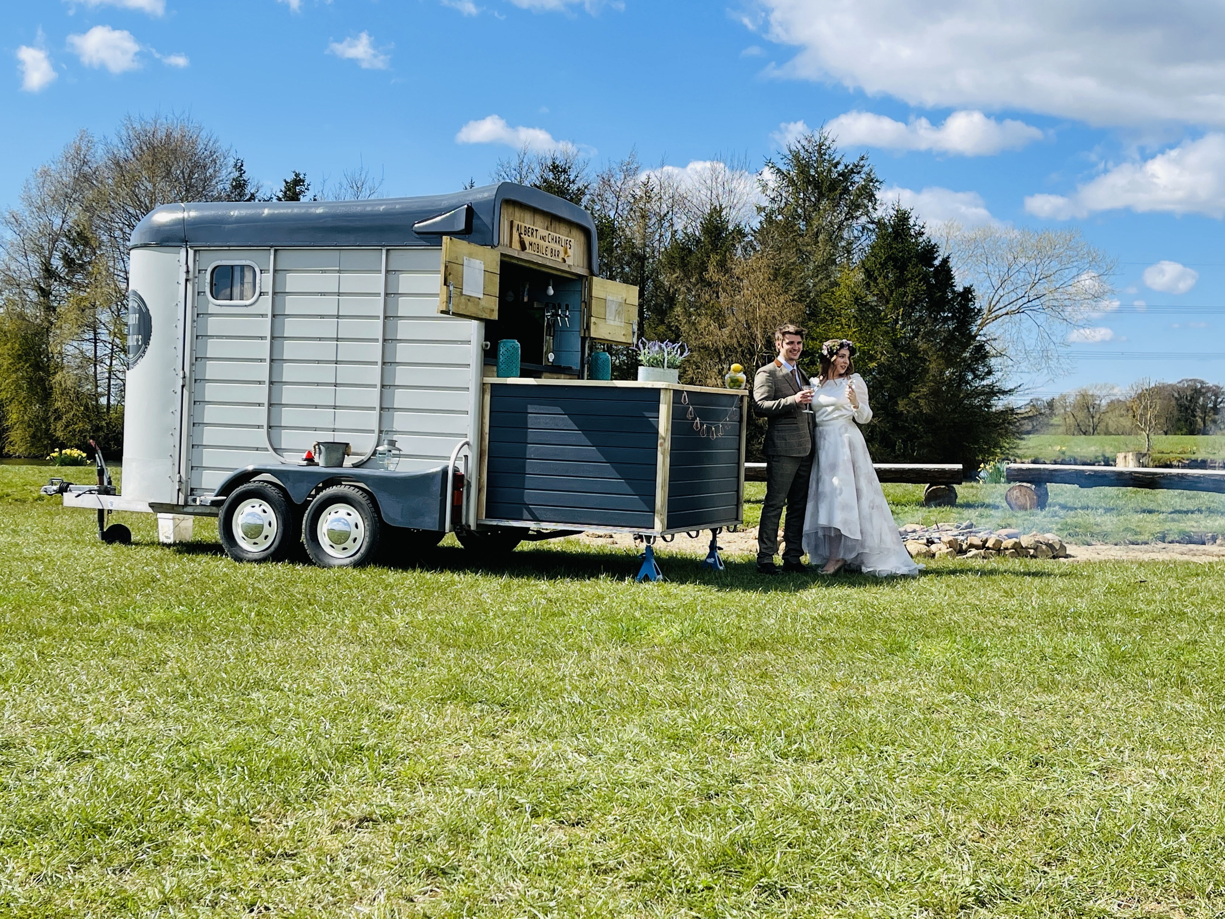 A charming mobile bar trailer sits on lush green grass, with a couple in formal attire raising glasses under bright, cheerful skies.