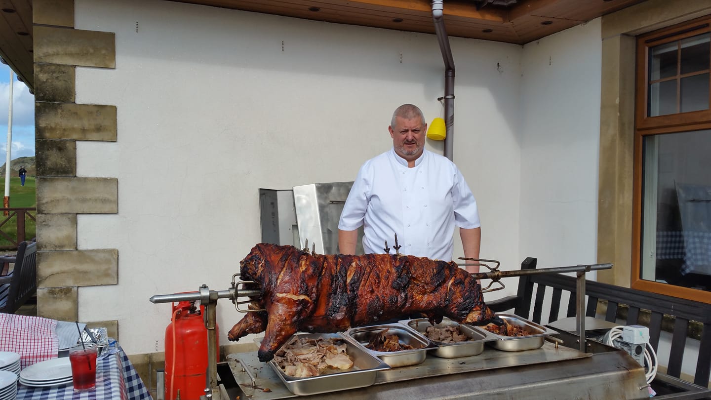 A chef stands proudly beside a beautifully roasted pig, adorned with crispy skin, under bright daylight, hinting at a quality outdoor catering setup.