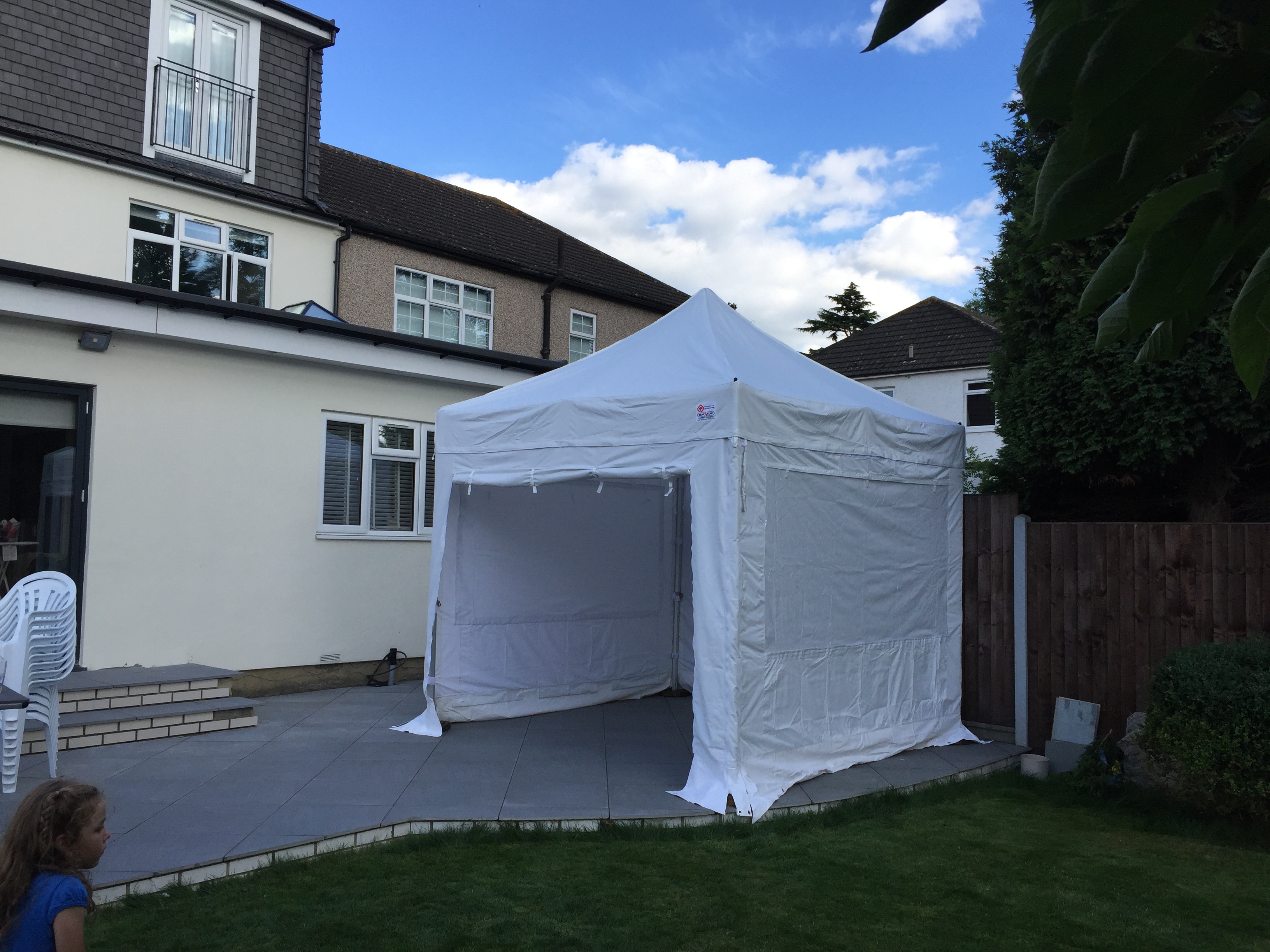 A white pop-up tent stands in a green lawn, surrounded by neat slate tiles, with a cozy outdoor atmosphere under a bright blue sky.