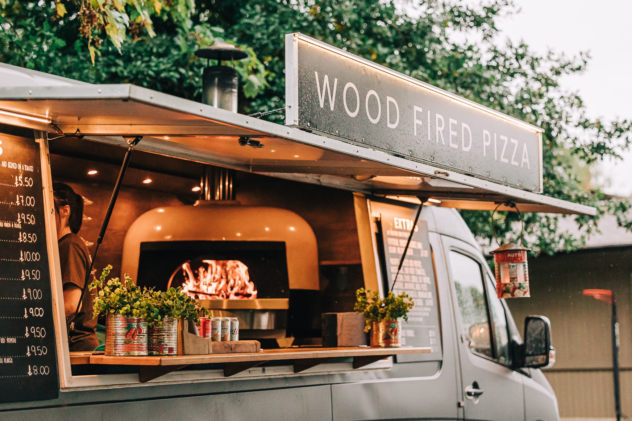 A cozy wood-fired pizza truck showcases a glowing oven and fresh herbs in vibrant cans. Warm lighting invites a friendly atmosphere.