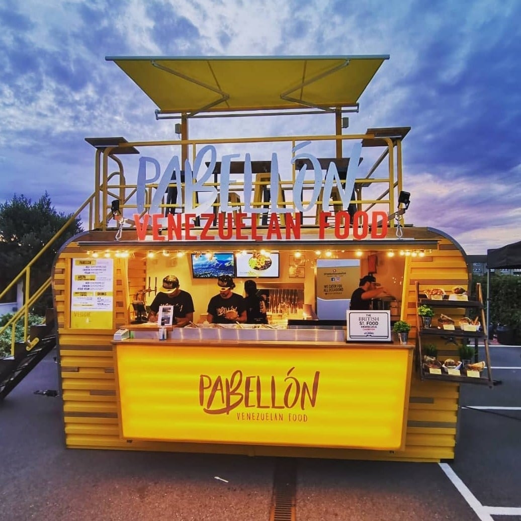 A vibrant yellow food truck, illuminated by warm lights, features chefs serving Venezuelan dishes, reflecting quality and authenticity.