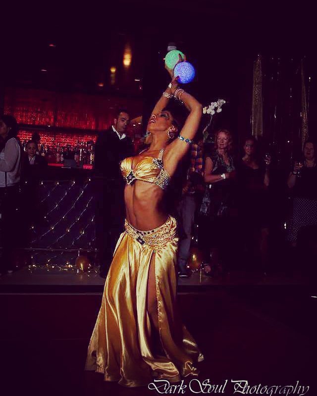 A spirited dancer in a shimmering gold costume captivates the crowd, illuminated by soft lighting in an elegant bar setting.