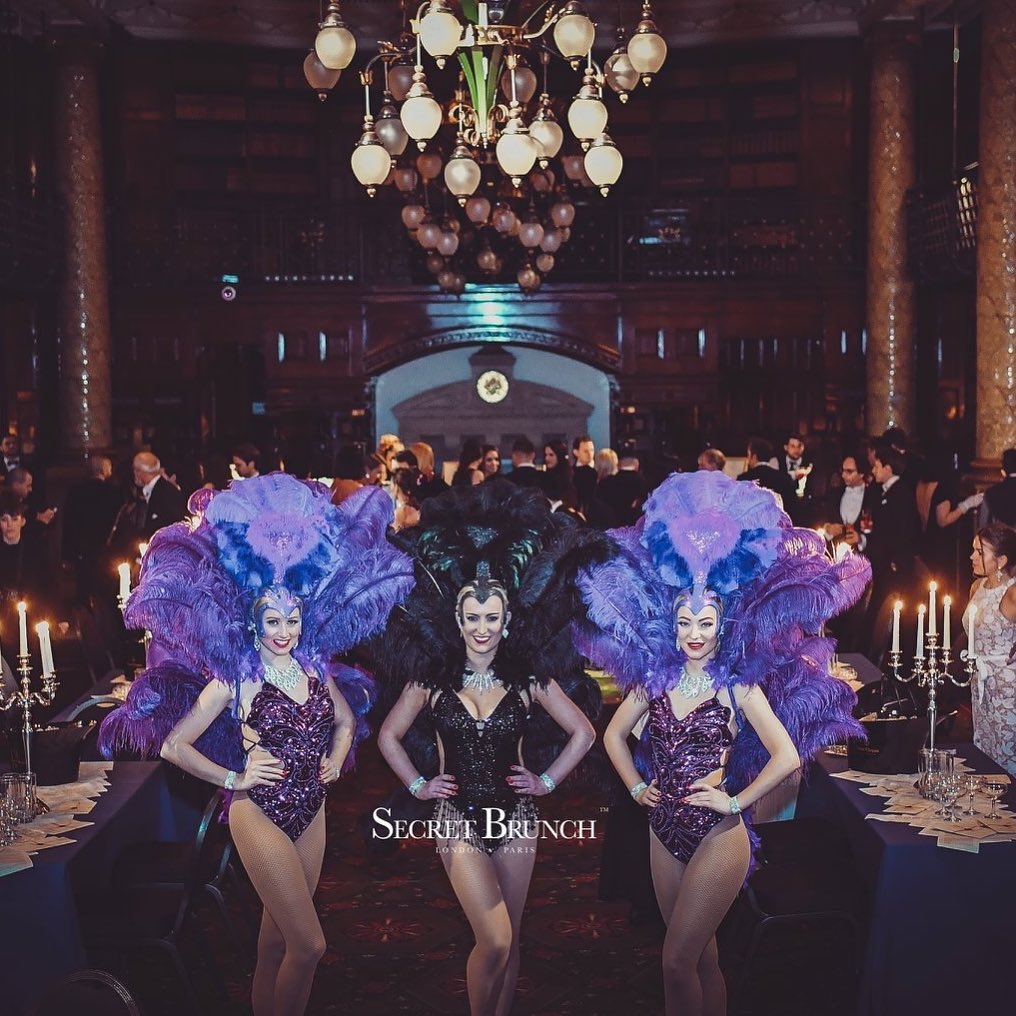 Colorful performers in sequined costumes and feather headdresses stand before elegantly dressed guests in a lavish, candlelit venue.