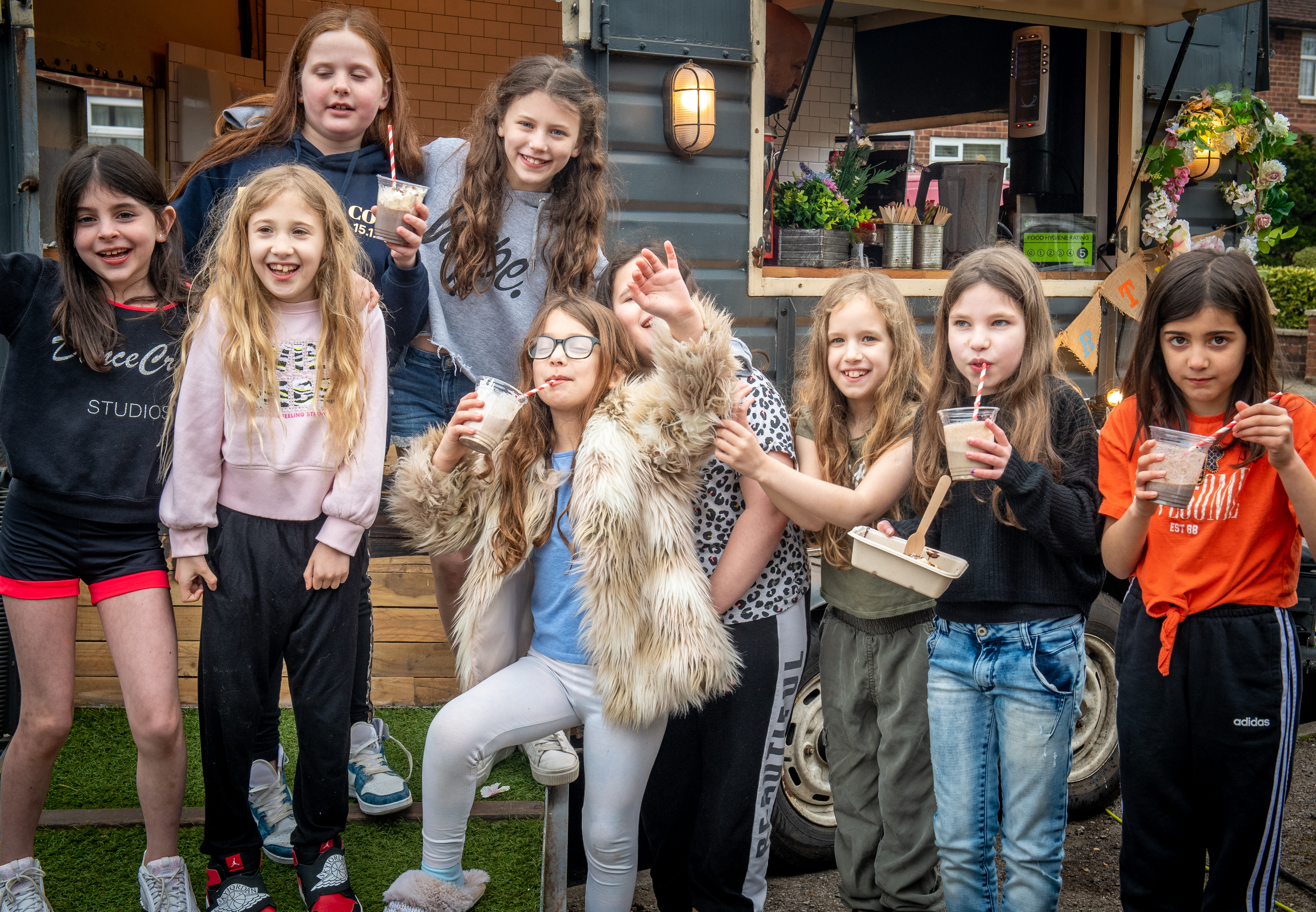 A cheerful group of girls gathers near a vintage food truck, enjoying milkshakes in clear cups, bright colors, and a lively atmosphere.