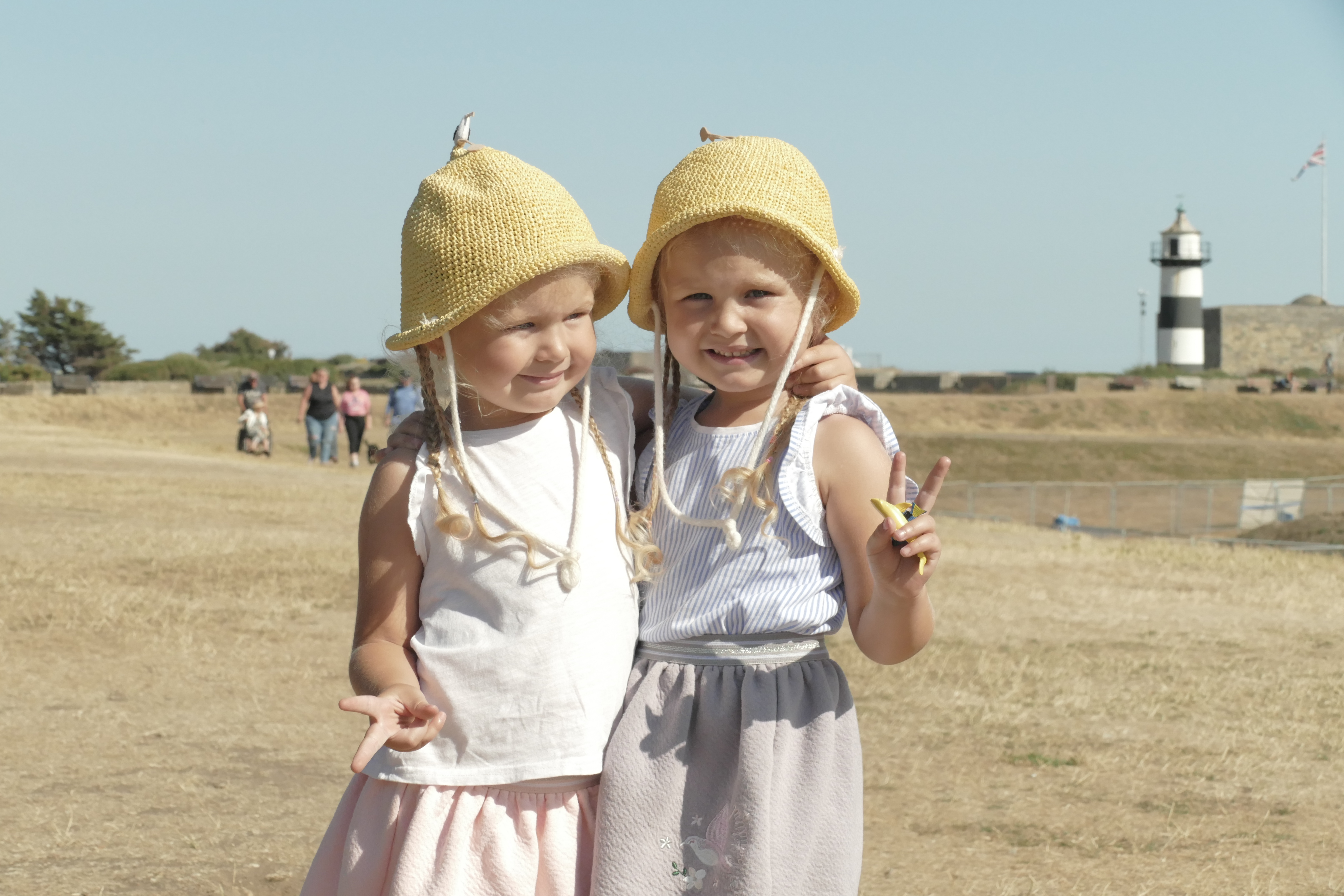 Two smiling girls in straw hats hug on a sunny field, with a black-and-white lighthouse behind them and bright, airy outdoor vibes.