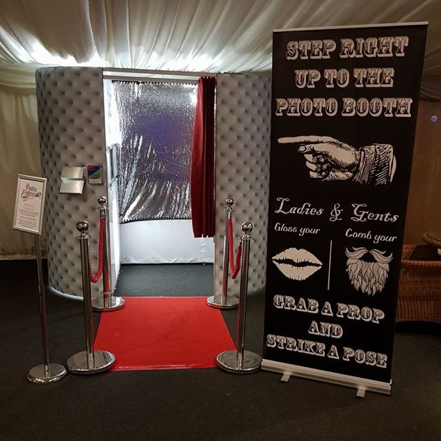 A chic photo booth entrance features a silver entrance, red carpet, and velvet ropes, exuding fun vibes in a warmly lit setting.