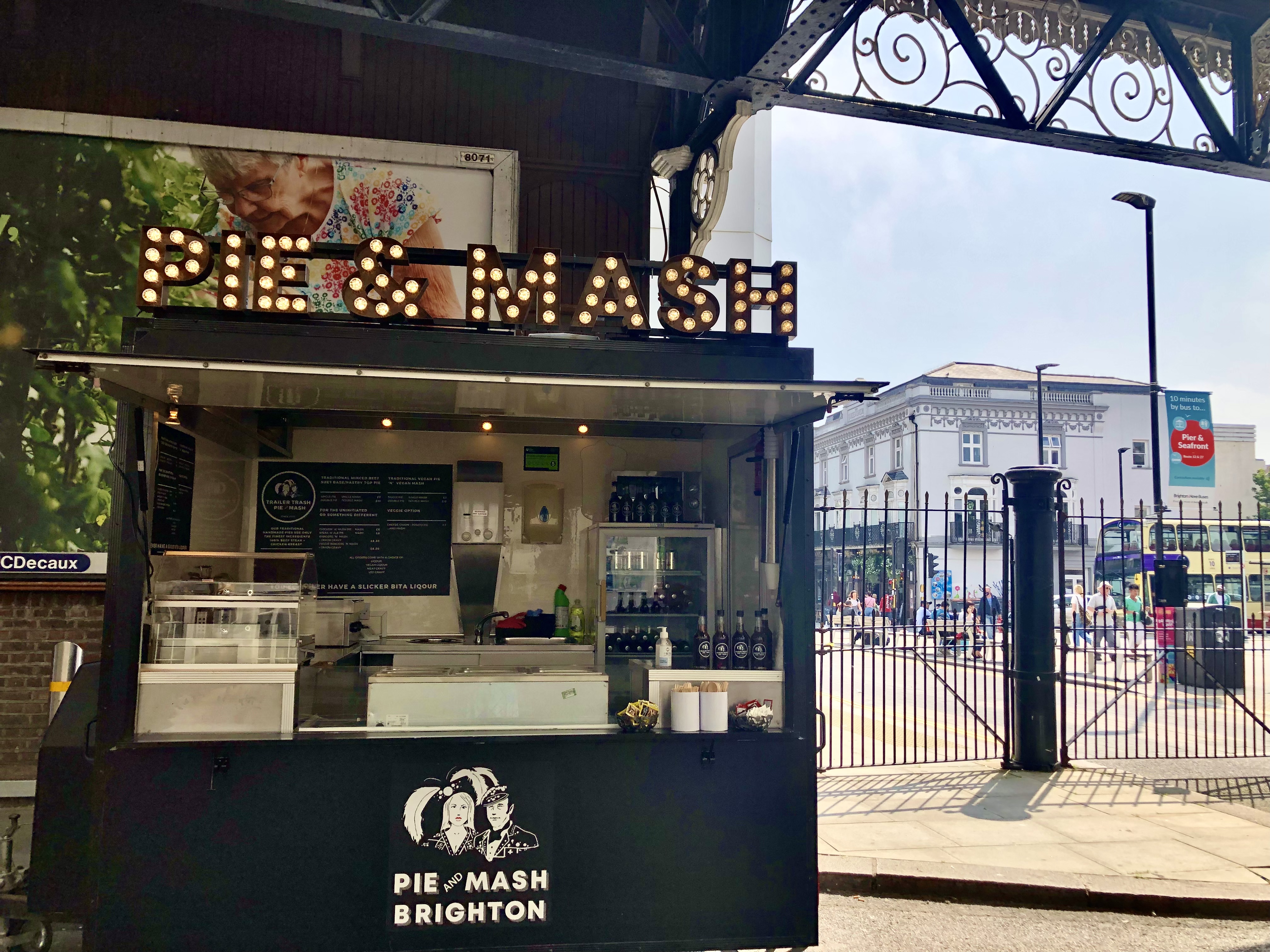 A sleek black food truck with a bright "PIE & MASH" sign stands against a bustling city backdrop, highlighting casual outdoor dining vibes.