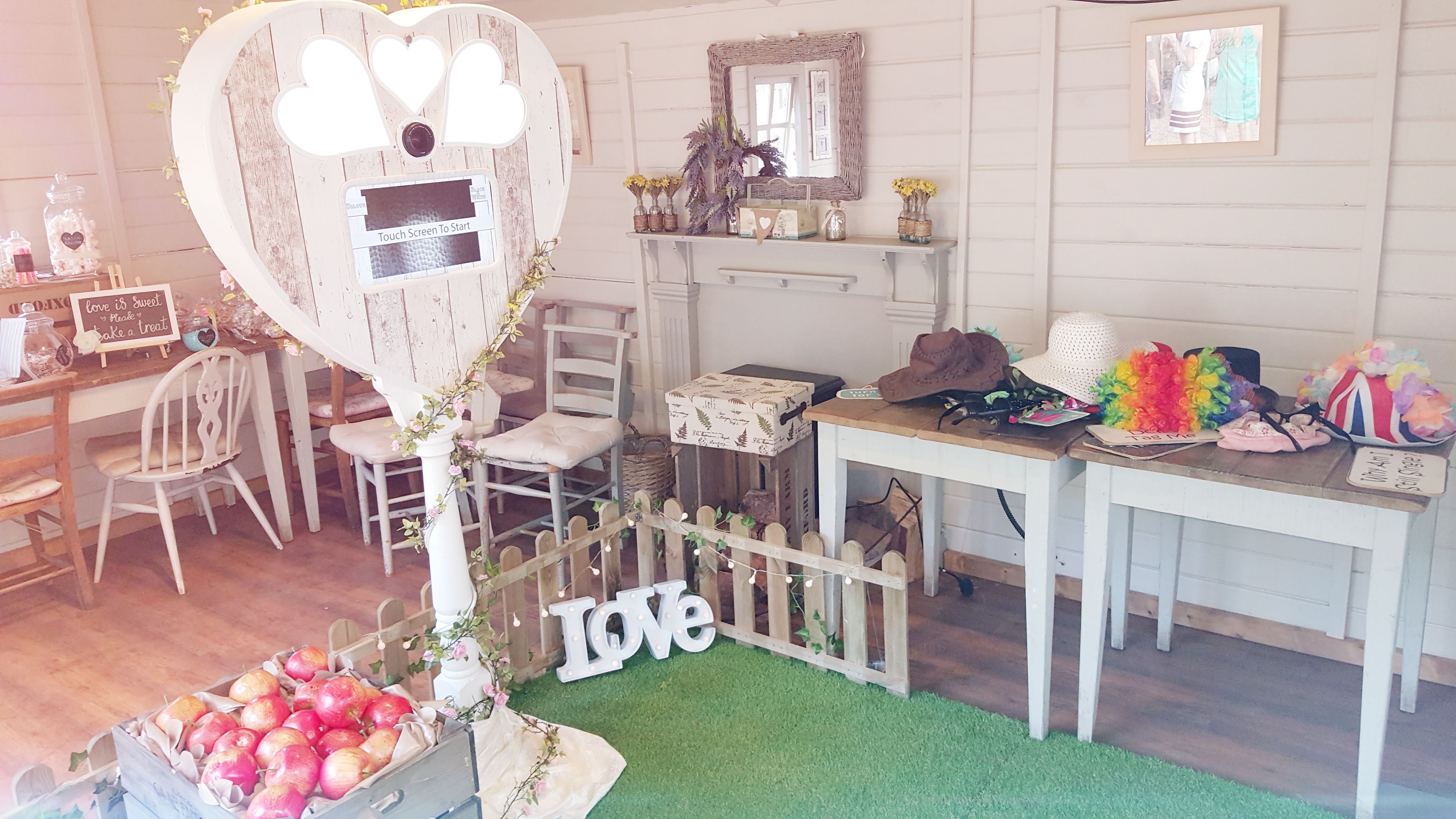 A cozy, light-filled space features a heart-shaped photo booth, rustic wooden tables with hats and colorful props, and warm wooden flooring.